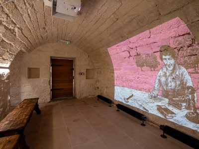 Historic stone room with a vaulted ceiling, where a projector displays an illustration on the wall. The projection depicts a woman at a desk with books and writing tools Historic stone room with a vaulted ceiling, where a projector displays an illustration on the wall. The projection depicts a woman at a desk with books and writing tools