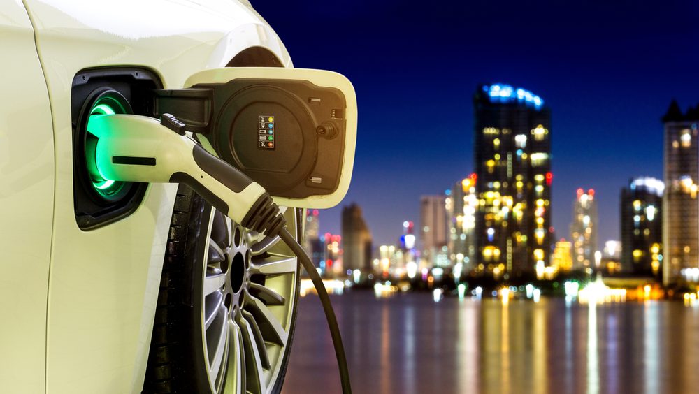 Electric vehicle at charging station with power cable supply plugged in with cityscape in background